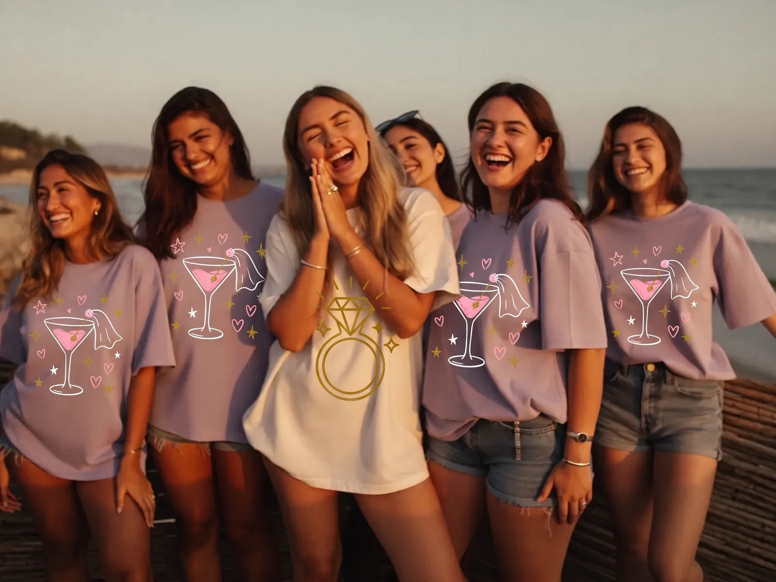 A bride in a cream t-shirt with a gold ring print laughing with her friends in lavender oversized t-shirts printed with a veiled cocktail glass, on a beach at sunset