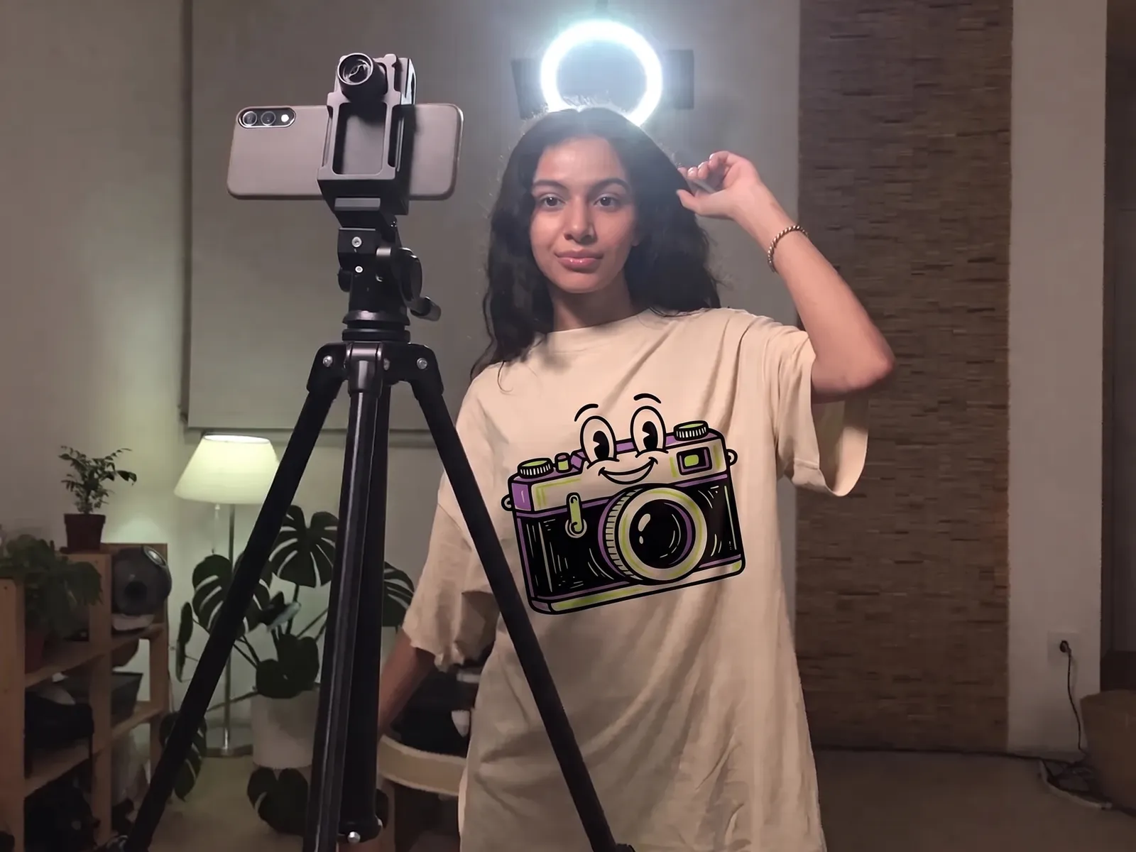 A young woman in a cream oversized t-shirt printed with a smiling cartoon camera, filming in front of a phone on a tripod and a ring light